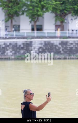 Vertikale Ansicht des Touristenfilms oder der Fotografie der kaukasischen Frau über das Telefon am Fluss Singapur in der Innenstadt. Genießen Sie die Stadtlandschaft um sie herum. Stockfoto