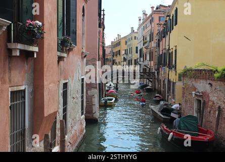 Ein venezianischer Kanal ist gesäumt von farbenfrohen historischen Gebäuden entlang des ruhigen Wassers. Die Boote liegen am Kanal, während Kajakfahrer unter einer Brücke gleiten. Stockfoto