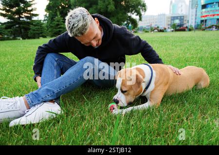 Hund mit einem Ball auf dem grünen Gras, ein Mann sitzt daneben. Der Besitzer streichelt den auf dem Gras liegenden Hund mit dem Ball. Ausbildung Amerikaner b Stockfoto