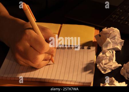 Eine Nahaufnahme einer Handschrift in einem Spiralnotizbuch unter warmer Beleuchtung. Die Szene umfasst zerknittertes Papier, Haftnotizen und einen Laptop, der auf Kreation hindeutet Stockfoto