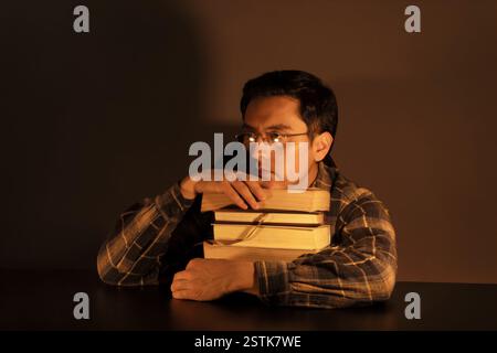Ein besinnlicher junger Mann in Brille und kariertem Hemd stützt sein Kinn auf gestapelten Büchern. Dunkles, warmes Licht betont seinen introspektiven Ausdruck, der ihn hervorruft Stockfoto