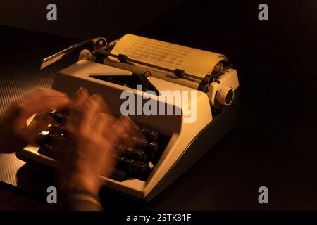 Verschwommene Hände, die auf einer weißen Retro-Schreibmaschine in einer düsteren, stimmungsvollen Umgebung tippen. Gelbes Papier mit eingetipptem Text erinnert an Kreativität und Zeit Stockfoto