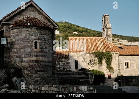 Antike Steinkirche mit roten Ziegeldächern vor einem grünen Hügel. Die verwitterte Architektur, die bogenförmigen Fenster und die mit Efeu bedeckten Wände. Altstadt B Stockfoto