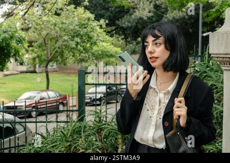 Eine junge Geschäftsfrau, die eine Sprachnachricht auf ihrem Smartphone sendet, während sie in der Stadt spaziert Stockfoto