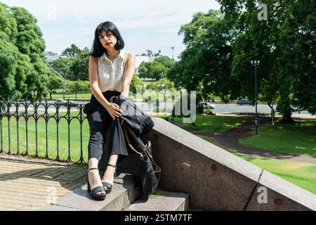 Geschäftsfrau sitzt auf Stufen in einem Park, hält Jacke und Tasche, genießt einen Moment der Ruhe Stockfoto