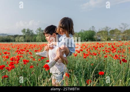 Seitenansicht des Huckepackreits eines jüngeren Mädchens auf einem älteren Kind in einem Mohnfeld Stockfoto