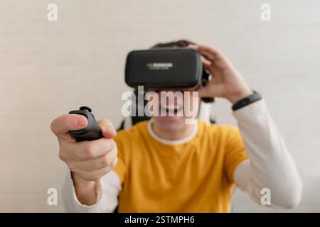 Junger Mann in VR-Brille, der Videospiel mit Joystick spielt, auf der Couch sitzt und dabei Virtual-Reality-Erfahrung hat Stockfoto