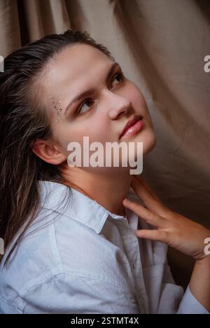 Nachdenkliche junge Frau mit Edelsteinen im Gesicht in entspannter Pose. Kaukasische Frau mit Glitzer im Gesicht. Beigefarbener Hintergrund. Stockfoto