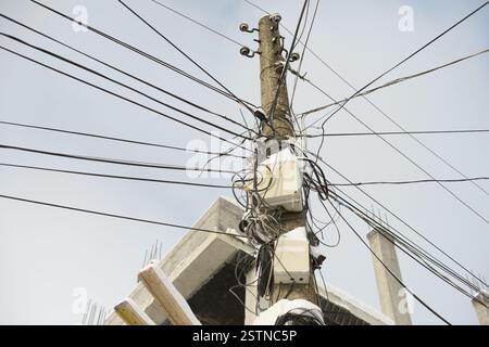 Ein verwitterter Beton-Versorgungsmast, der mit verwickelten elektrischen Drähten, Netzwerkkabeln und Anschlusskästen überlastet ist, vor einem blauen Himmel in der Nähe einer Konstruktion Stockfoto