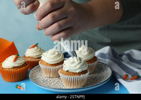 Frau quetscht Schlagsahne auf köstlichen Karotten-Cupcakes an blauem Tisch, Nahaufnahme Stockfoto