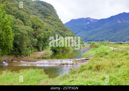 Goesan County, Südkorea - 10. September 2020: Ein kleines Wehr stürzt sanft über einen ländlichen Fluss, umgeben von üppigem Grün, mit Bergen in der Stockfoto
