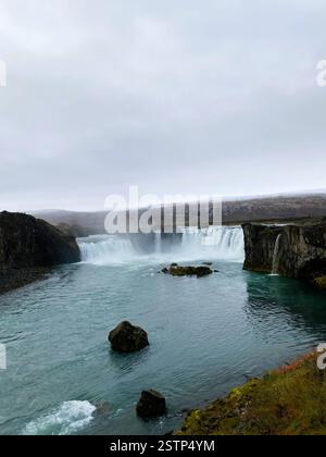 Das Bild fängt eine atemberaubende Wasserfallszene ein, die über felsige Felsvorsprünge in einen ruhigen türkisfarbenen Fluss stürzt, umgeben von zerklüfteten isländischen Landschaften Stockfoto