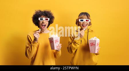 Aufgeregter Mann und Frau gelbe Sweatshirts mit 3D-Kino-Brille vor einem gelben Hintergrund, der Popcorn hält Stockfoto