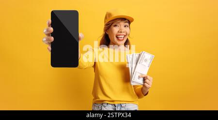Asiatische Frau, die Geld und ein Handy vor gelbem Hintergrund hält Stockfoto