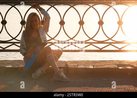 Glückliche Frau mit schickem Scateboard sitzt am Wasser in der Sonne. Schöne und modische junge Frau posiert mit einem Skateboa Stockfoto