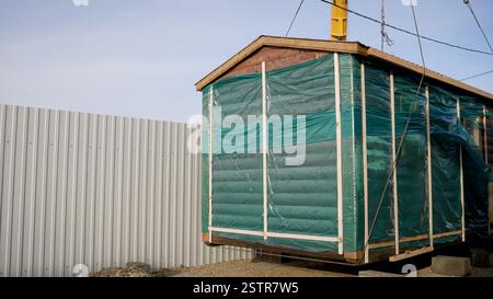 Vorgefertigtes Holzhaus-Modul zum Anheben von Kranen auf der Baustelle mit grüner Schutzfolie abgedeckt Stockfoto
