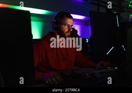 Junger Mann mit Brille und Kopfhörern, der in einem Computerspielzimmer sitzt und sich auf einen Bildschirm konzentriert, mit farbenfrohen Neonlichtern im Hintergrund. Stockfoto