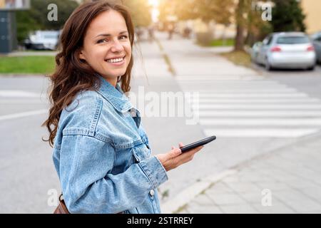 Lächelnde junge städtische Frau in lässigem Outfit, die die Straße entlang läuft, während sie ihr Smartphone hält Stockfoto