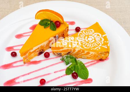 Zwei Scheiben Mango-Käsekuchen werden elegant auf einem Teller präsentiert, dekoriert mit Minzblättern und einer Beerensoße. Frische Orangenscheiben verbessern die vibra Stockfoto