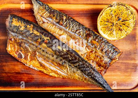 Zwei Portionen perfekt gegrillter Makrele werden auf einem Holzbrett präsentiert, zusammen mit einer angekohlten Zitronenhälfte. Das Gericht bietet ein reichhaltiges und appetitliches t Stockfoto