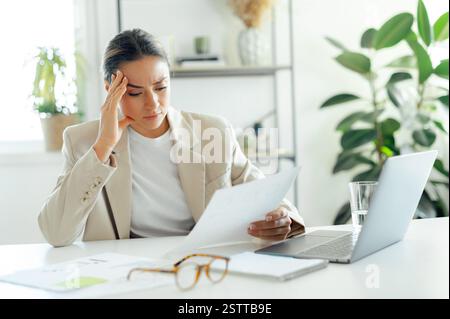 Frustrierte kaukasische Geschäftsfrau, die aufgrund der Arbeitsergebnisse gestresst aussieht, beim Lesen eines Dokuments an ihrem Schreibtisch den Kopf hält, verwirrt, verärgert und enttäuscht ist Stockfoto