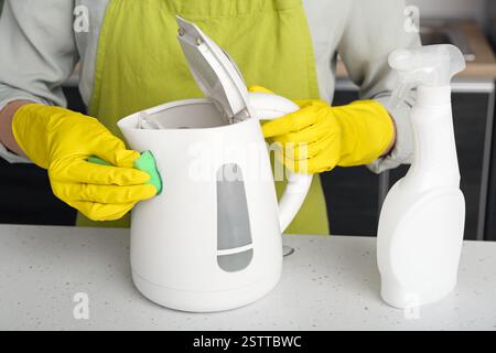 Frau in gelben Handschuhen, die elektrischen Wasserkocher auf einem weißen Tisch reinigt Stockfoto