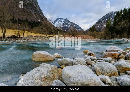 Schneebedeckte Berge stehen hoch vor einem blauen Himmel, während ein ruhiger Fluss über glatte Steine fließt und das Ende des Winters signalisiert. Stockfoto