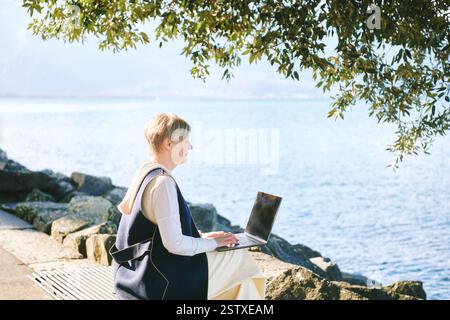 Außenporträt einer Reifen Frau mit Laptop, die auf der Bank neben dem See sitzt Stockfoto