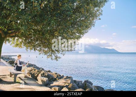 Landschaftsbild des Genfer Sees und der schweizer Alpen, reife Frau, die unter grünem Baum sitzt und mit Laptop arbeitet Stockfoto