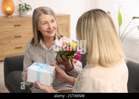 Lächelnde Mutter erhält einen Tulpenstrauß und ein Geschenk von ihrer Tochter auf dem Sofa zu Hause. Alles Gute zum Muttertag Stockfoto