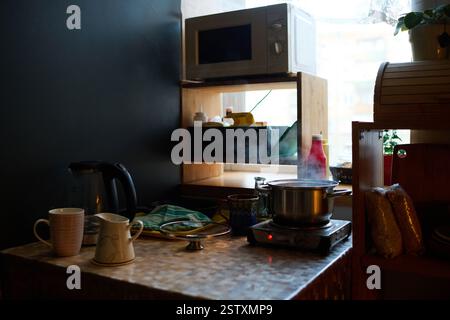 Küchenszene mit Mikrowelle auf Holzregal, Teekanne auf der Theke und Kochutensilien, sanftem Tageslicht, das gemütliche Interieur beleuchtet und eine gemütliche Atmosphäre schafft Stockfoto