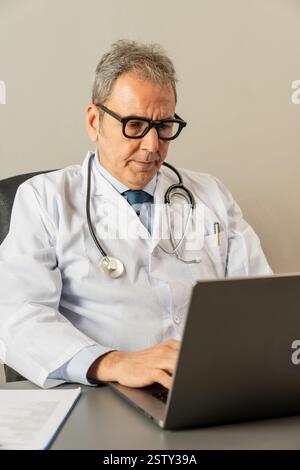Oberarzt mit Laptop in seiner Arztpraxis, konzentriert auf Arbeit und Online-Beratung. Stockfoto