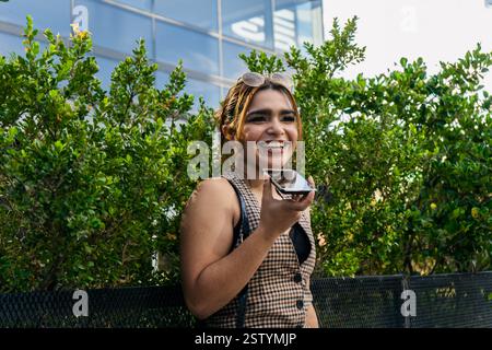 Eine junge Geschäftsfrau, die im Freien mit ihrem Smartphone eine Sprachnotiz sendet, lächelt und wegblickt Stockfoto