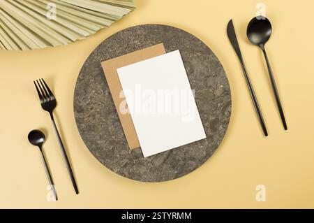 Blick von oben auf braunen Marmorteller, schwarzes Besteck, weiße und braune Karten auf gelbem Hintergrund. Getrocknete Palmblätter. Essenspräsentation, Steintablett. Stockfoto