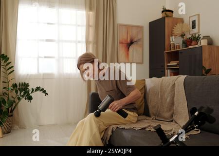 Ältere Frau, die auf einem bequemen Sofa in einem gut beleuchteten Wohnzimmer sitzt, umgeben von Pflanzen und modernem Dekor, während sie mit einem Gadget beschäftigt ist Stockfoto