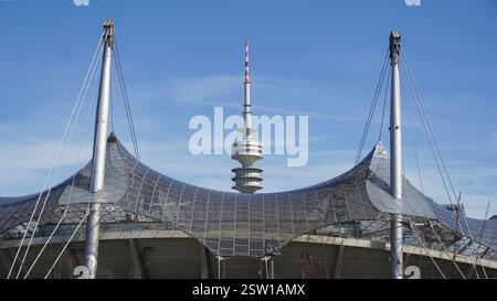 Foliendachkonstruktion und Tribüne des Olympiastadions in München mit dem hohen Olympiaturm im Hintergrund unter blauem Himmel bei Sonnenschein Stockfoto