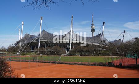 Berühmtes Olympiastadion in München mit Trainingsgelände im Vordergrund und dem hohen Olympiaturm im Hintergrund unter blauem Himmel bei Sonnenschein Stockfoto