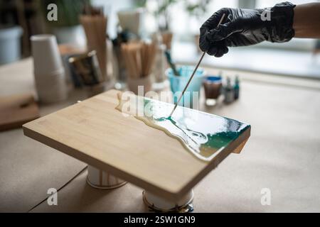 Handfläche einer künstlerischen, akribischen, aufmerksamen Frau in schwarzem Handschuh, die an Epoxidharzmalerei arbeitet. Stockfoto
