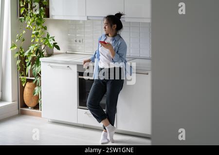 Besorgte Asiatin der Millennials, die in der Küche steht und ein Handy hält und auf einen wichtigen Anruf wartet Stockfoto