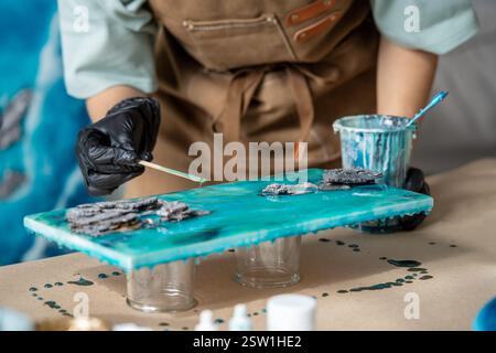 Die Künstlerin gießt Epoxidharz und schmiert es mit einem Stock auf Holzbrett, mit Hilfe von Flüssigkunsttechnik Stockfoto