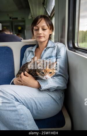 Nachdenkliche Frau, die sich umarmt und mit einer Katze im Zug reist. Gestresste Kätzchen, die auf den Schoßen der Frau sitzen Stockfoto