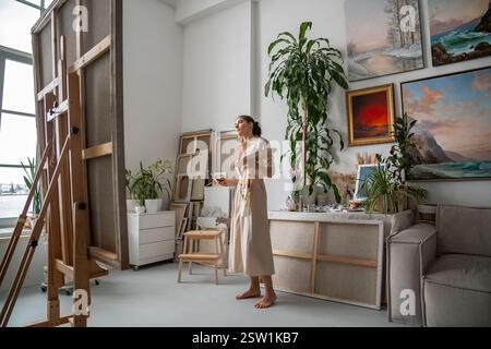 Eine nachdenkliche Künstlerin mit einer Palette von Ölfarben und Pinseln steht vor der Staffelei mit Leinwand. Stockfoto