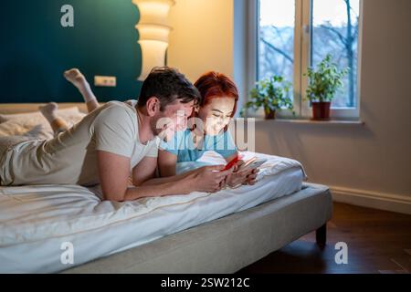 Glückliches Paar, Frau, Ehemann im Bett, schaute in das Smartphone, lächelte, unterhielt sich im Bett. Stockfoto