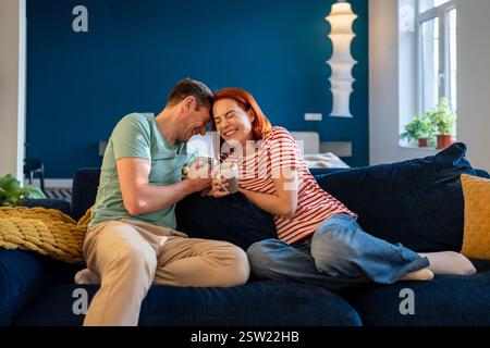 Glückliches, sorgenfreies, junges Paar, das sich verliebte und lachte, während es sich auf der Couch mit Kaffee entspannte Stockfoto