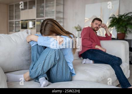 Eine Frau in Tränen, Traurigkeit, versteckt das Gesicht, schrumpft auf dem Sofa, während ihr verzweifelter Ehemann am anderen Ende sitzt Stockfoto