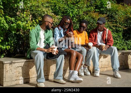 Entspannte Gruppe afroamerikanischer Freunde, die auf der Parkbank sitzen und Kaffee trinken, während sie sich unterhalten Stockfoto