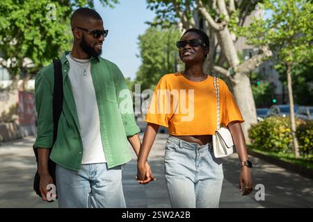 Fröhliches afroamerikanisches Liebespaar, das in der Stadt Hände hält, bringt Freundin zum Lachen. Stockfoto