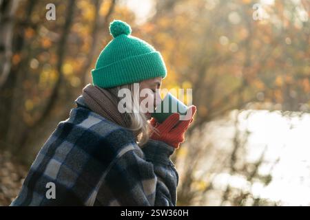 Eine ruhige Frau schlürft heißen Tee aus der Thermoskanne im Herbstwald am See. Langsames Leben auf Alleinreisen Stockfoto