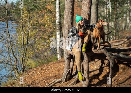 Sorglose Frau, die mit Hund im Herbstwald geht, genießt heiße Thermos-Tee-Pause auf Baumwurzeln in der Nähe des Flusses. Stockfoto