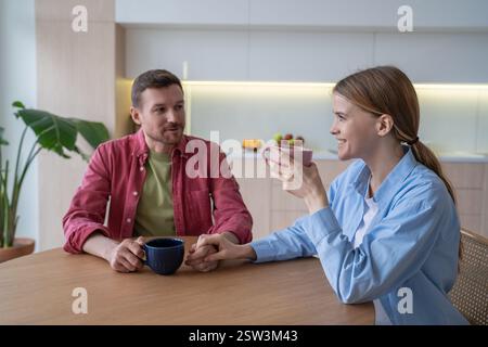 Lächelndes junges Paar, das in die Ferne starrt, während es zusammen Tee und Frühstück in der Küche hatte Stockfoto